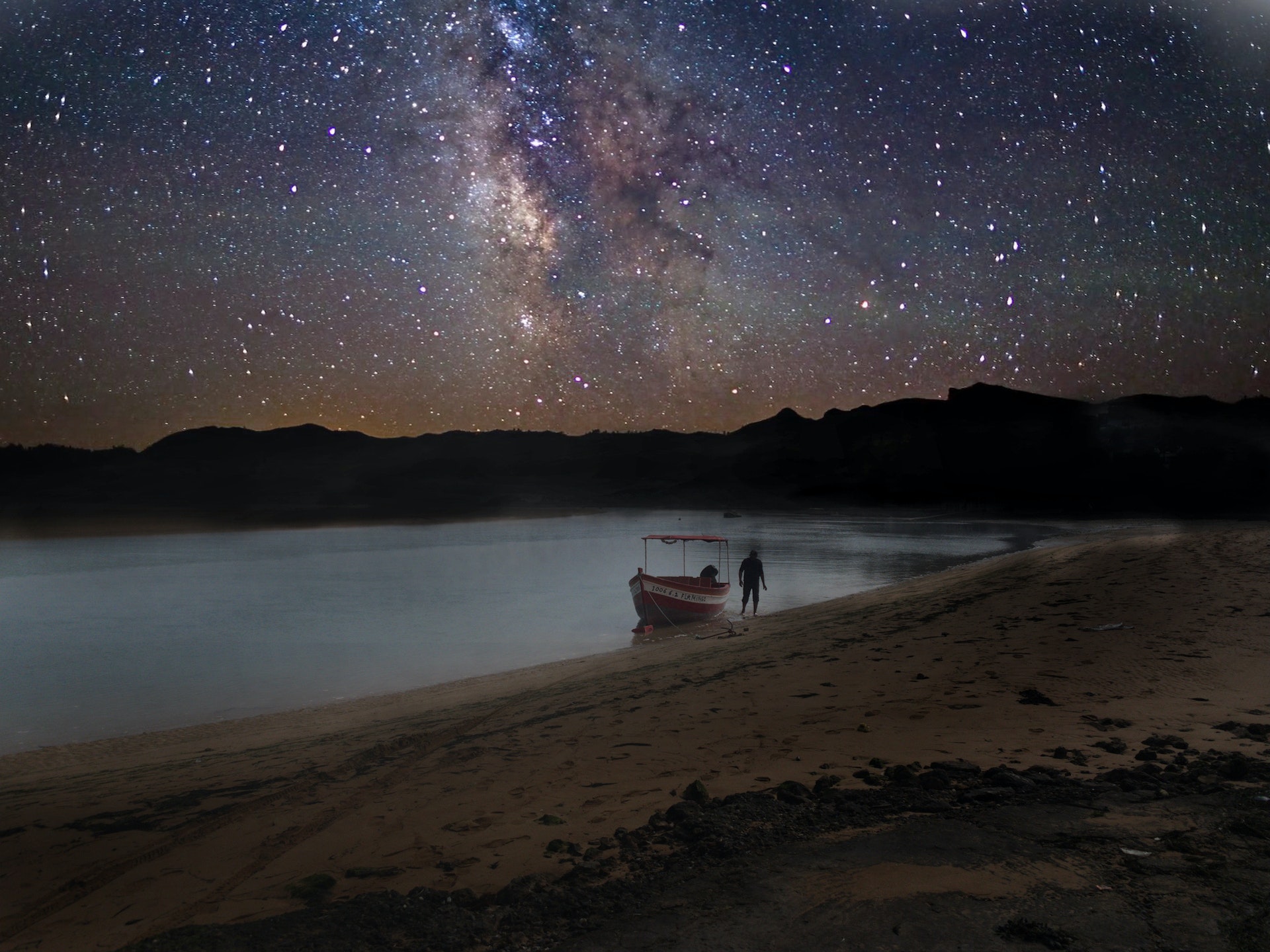 Sous une couverture d'étoiles scintillantes la nuit, un homme marche près d'un bateau de pêche qui flotte sur la mer à Oualidia au Maroc