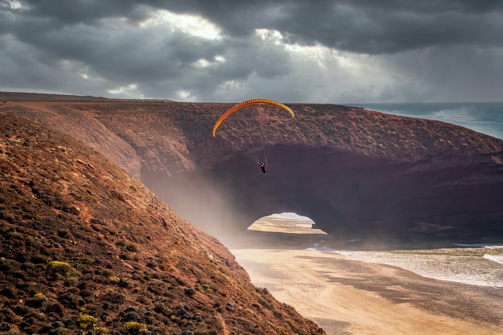 Un parapente tombe sur le sable rouge à côté de l'arche terrestre de la plage de Legzira, au Maroc au coucher du soleil alors que le vent fouette les grains de sable